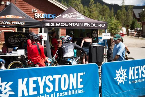 Image of a bike event at Keystone, featuring a large tent labeled "BIG MOUNTAIN ENDURO" and individuals interacting at an information booth. There are several cyclists in riding gear, along with spectators near a fence displaying the “KEYSTONE” logo. The backdrop includes trees and mountains under a clear blue sky. Keystone Resort Bike Park mountain bike trail.