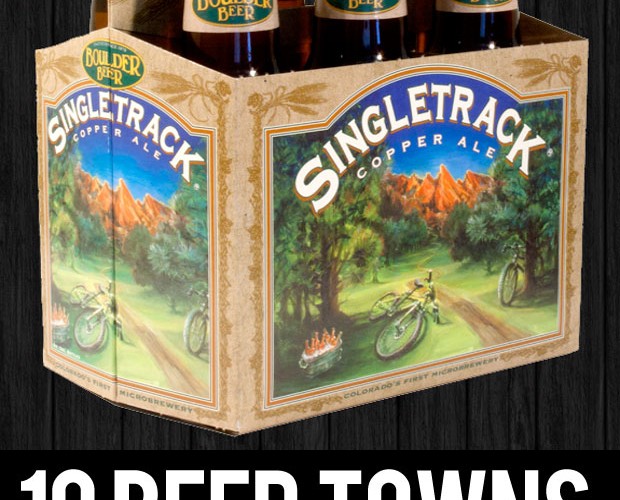 A six-pack of Boulder Beer’s Singletrack Copper Ale, featuring an illustrated label with a mountain biking scene surrounded by lush greenery and mountains, set against a dark wood background. The text below reads, "10 BEER TOWNS with a mountain biking problem."