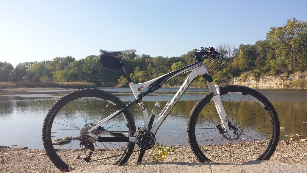Scott Spark 29 Team: A white mountain bike positioned beside a tranquil riverbank, surrounded by lush green trees and a clear blue sky. The bike features robust tires and a sleek design, with the water reflecting the natural scenery in the background.