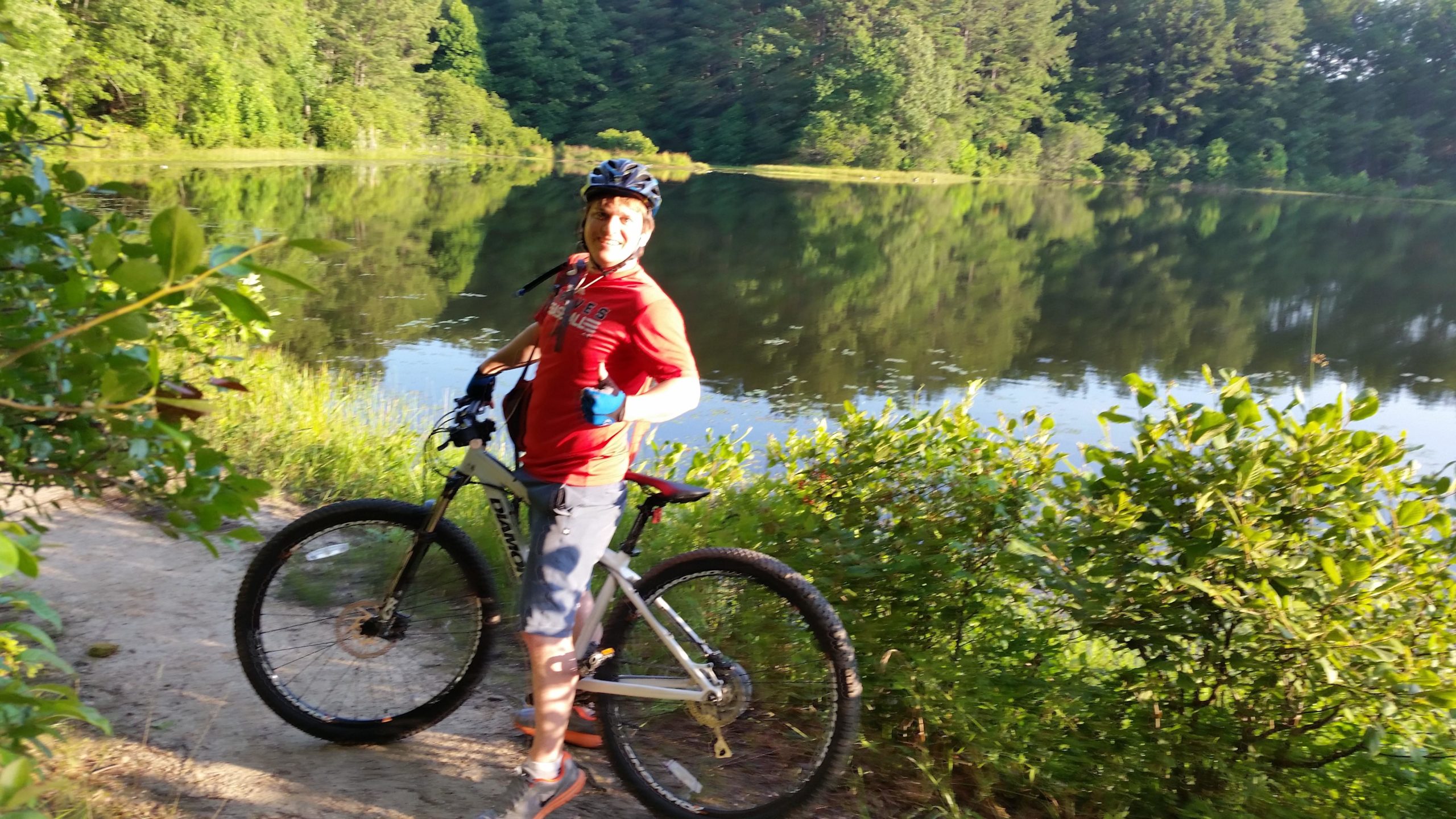 A person standing next to a mountain bike on a dirt path beside a tranquil lake, surrounded by lush green foliage and trees. The individual is smiling and giving a thumbs-up, wearing a red shirt and a black helmet. The calm water reflects the greenery, creating a serene outdoor atmosphere. Tribble Mill Park mountain bike trail.