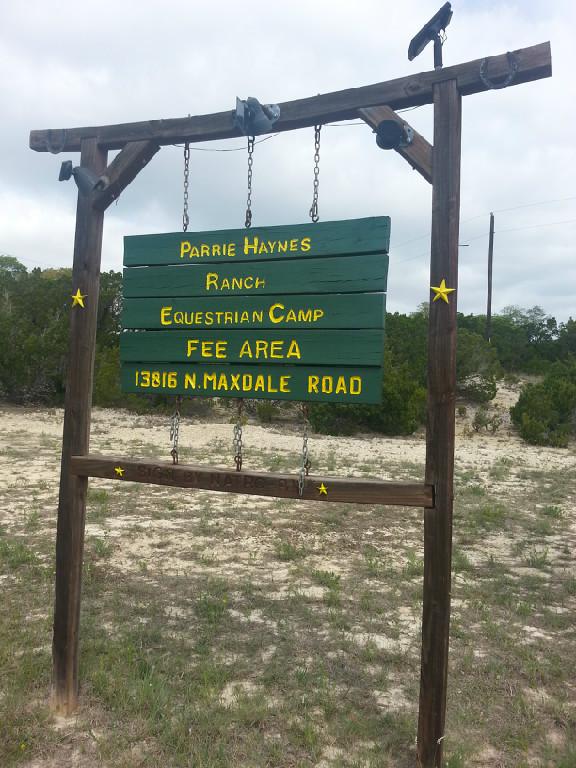A wooden sign reading "Parrie Haynes Ranch Equestrian Camp Fee Area" hanging from a frame, with the address "13816 N. Maxdale Road" below. The sign features yellow lettering and decorative stars, set against a natural landscape with low shrubs and a cloudy sky. Parrie Haynes Ranch Equestrian Center mountain bike trail.