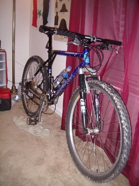 GT Timberline: Mountain bike parked indoors, featuring a black and blue frame, with water bottles mounted on the frame. The bike rests on a carpeted floor next to a window with red curtains, and a small stack of clothes is visible nearby. A storage unit can be seen in the background, along with some wall decorations.