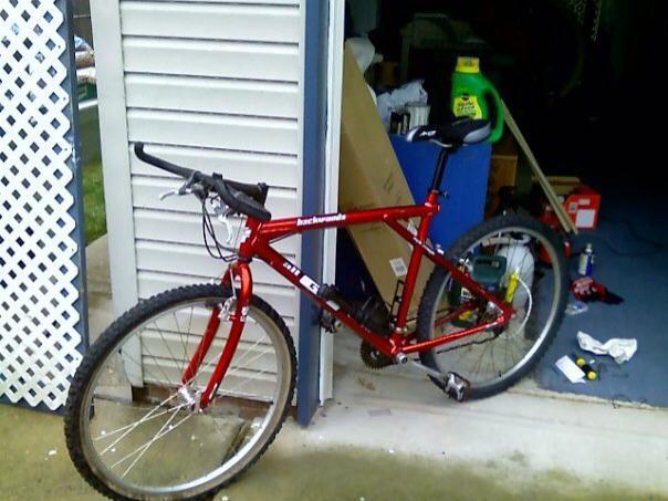 GT Backwoods: A red mountain bike parked near a garage entrance, with various items and boxes visible in the background. The bike features thick tires and a sturdy frame, indicating it is intended for off-road use.
