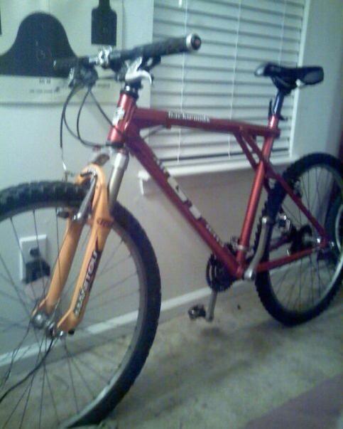 GT Backwoods: Mountain bike leaning against a wall, featuring a red frame and bright orange front suspension fork, with visible handlebars and two wheels on a carpeted floor. In the background, there are window blinds and a partially visible wall.