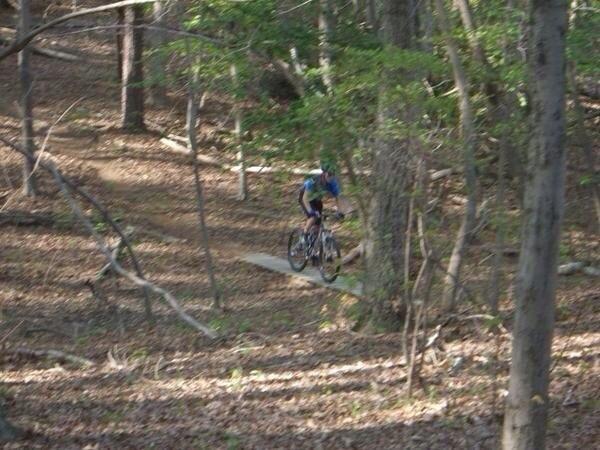 Giant NRS 2: A cyclist riding along a narrow trail through a wooded area, surrounded by trees and fallen leaves. The cyclist is wearing a blue and black outfit and is navigating a section of the path.