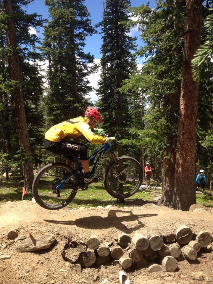 A mountain biker in a yellow jacket and red helmet jumps over a wooden log obstacle in a forested area, surrounded by tall pine trees. Other bikers can be seen in the background, enjoying the trail on a sunny day with blue skies. Keystone Resort Bike Park mountain bike trail.