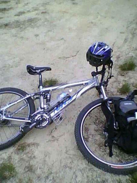 Giant NRS 2: A silver mountain bike rests on a gravel path, next to a black backpack and a blue helmet positioned on the bike's handlebars. The surroundings appear natural and earthy, suggesting an outdoor adventure or cycling environment.