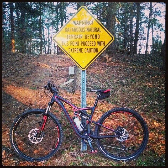 Specialized Camber 29: A mountain bike is leaning against a yellow warning sign that reads, "WARNING! HAZARDOUS NATURAL TERRAIN BEYOND THIS POINT PROCEED WITH EXTREME CAUTION." The bike has black and red detailing and is situated on a trail surrounded by trees and fallen leaves.
