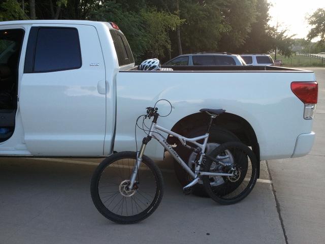 Gary Fisher HiFi: A silver mountain bike is leaning against the back of a white pickup truck parked in a driveway. A white bicycle helmet sits on the truck's bed, and greenery can be seen in the background.