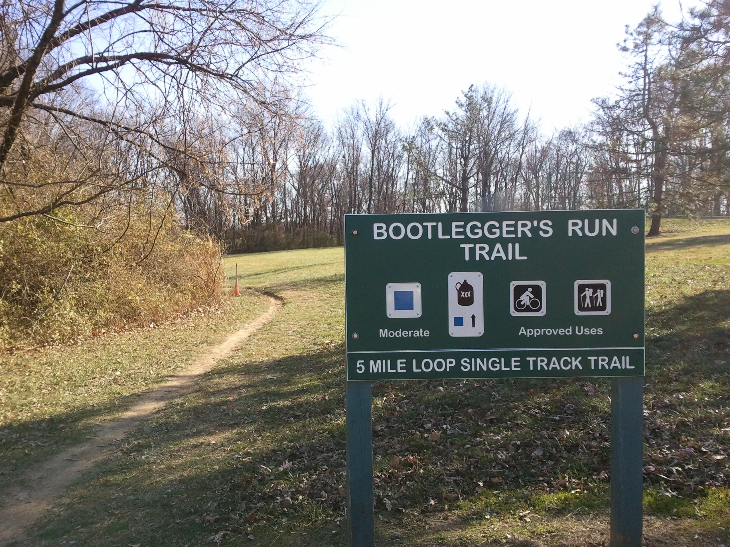 Sign for Bootlegger's Run Trail indicating a moderate difficulty level, approved uses for biking and hiking, and describing the trail as a 5-mile loop single track trail. The background features a grassy area and bare trees under a clear sky. Creve Couer Park mountain bike trail.