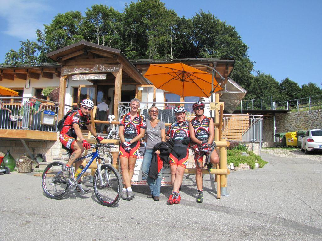Group of cyclists in athletic gear, posing outside a mountain bar named "Bar Zattron al Passo." The scene features three cyclists with bikes and a woman smiling in the center. The bar has a wooden exterior with an orange umbrella, surrounded by greenery. Bright sunny day, conveying a cheerful atmosphere. San Giovanni Al Monte - Lake Garda mountain bike trail.