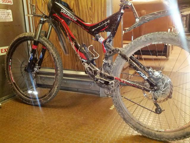 Specialized Stumpjumper FSR: A close-up view of a black and red mountain bike leaning against a doorway, featuring dirty tires and a muddy frame, indicative of recent use on rugged terrain. The interior of the space has wooden paneling and visible seating in the background.
