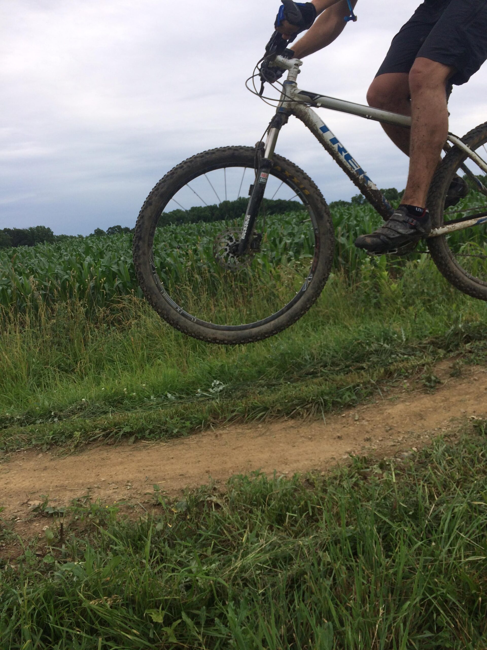 Trek Mamba: A mountain biker is captured in mid-air while jumping off a dirt path, with a field of corn in the background under a cloudy sky. The biker wears shorts and gloves, and the mountain bike shows dirt and mud, indicating off-road use.