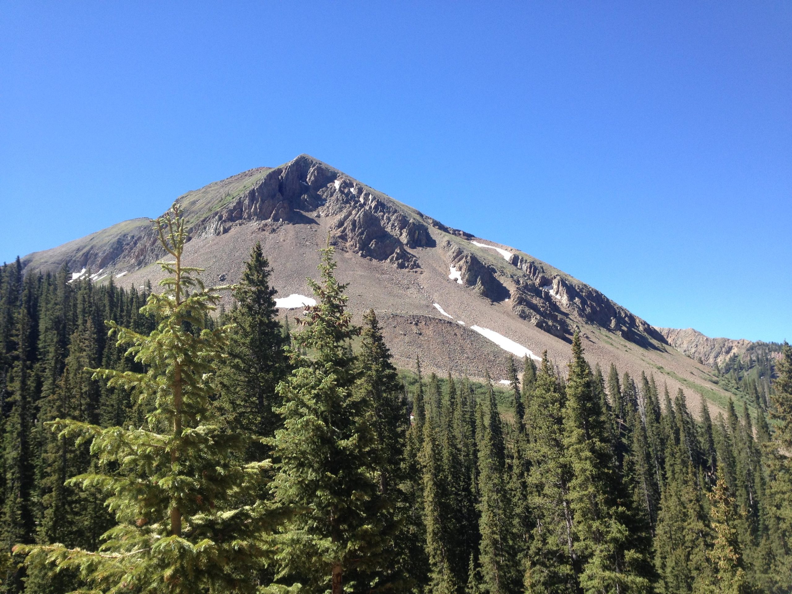 A scenic view of a mountain peak rising above a dense forest of evergreen trees, under a clear blue sky. The landscape features rocky terrain and patches of snow on the mountain, creating a contrast with the greenery below. Colorado Trail: Kennebec Pass To Junction Creek mountain bike trail.