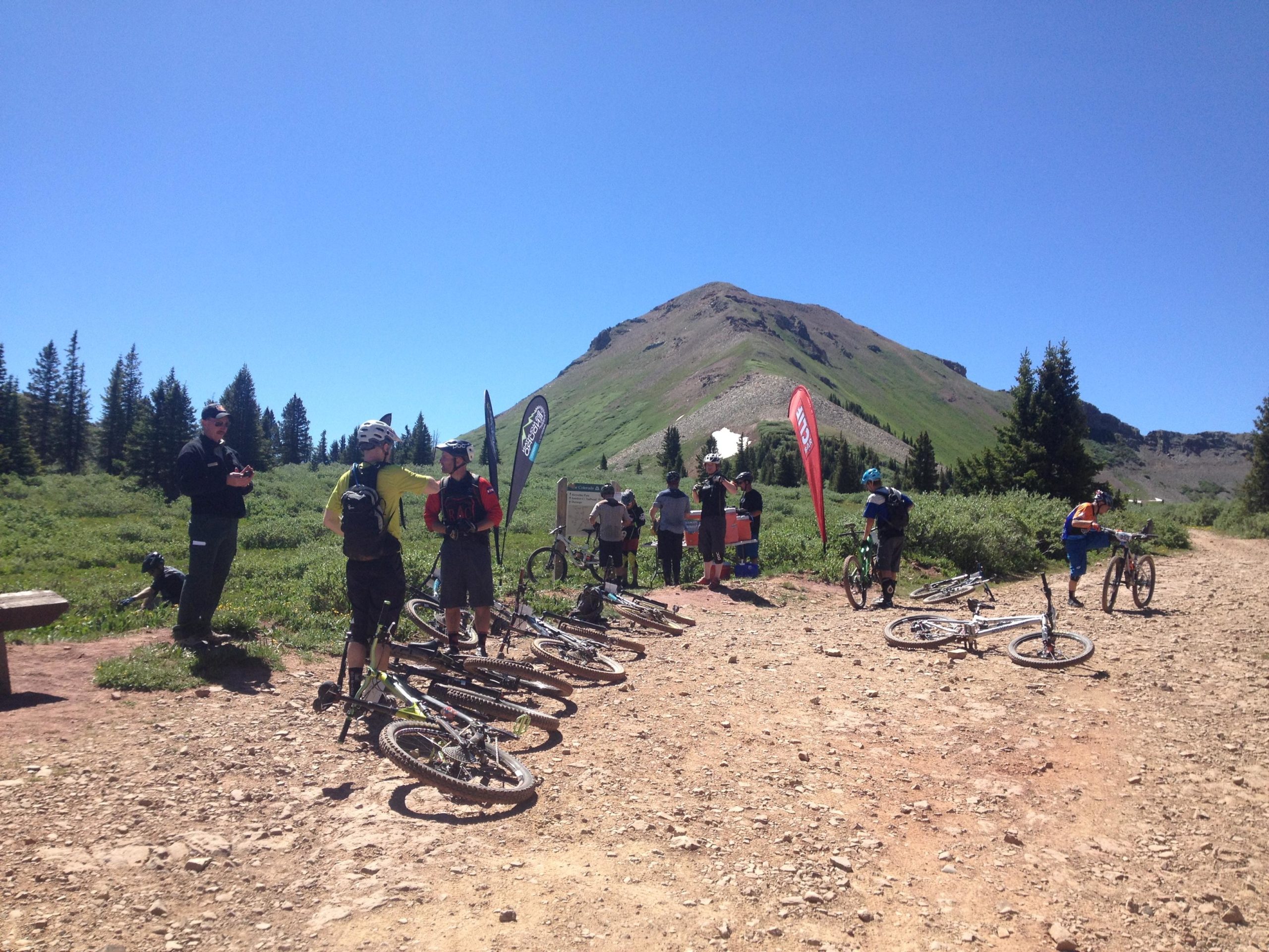 A group of mountain bikers gathers near a dirt trail surrounded by greenery and trees, with a mountain in the background. Some riders stand conversing, while others adjust their bikes. Flags are visible in the scene, indicating a biking event or gathering. The sky is clear and blue, suggesting a sunny day. Colorado Trail: Kennebec Pass To Junction Creek mountain bike trail.