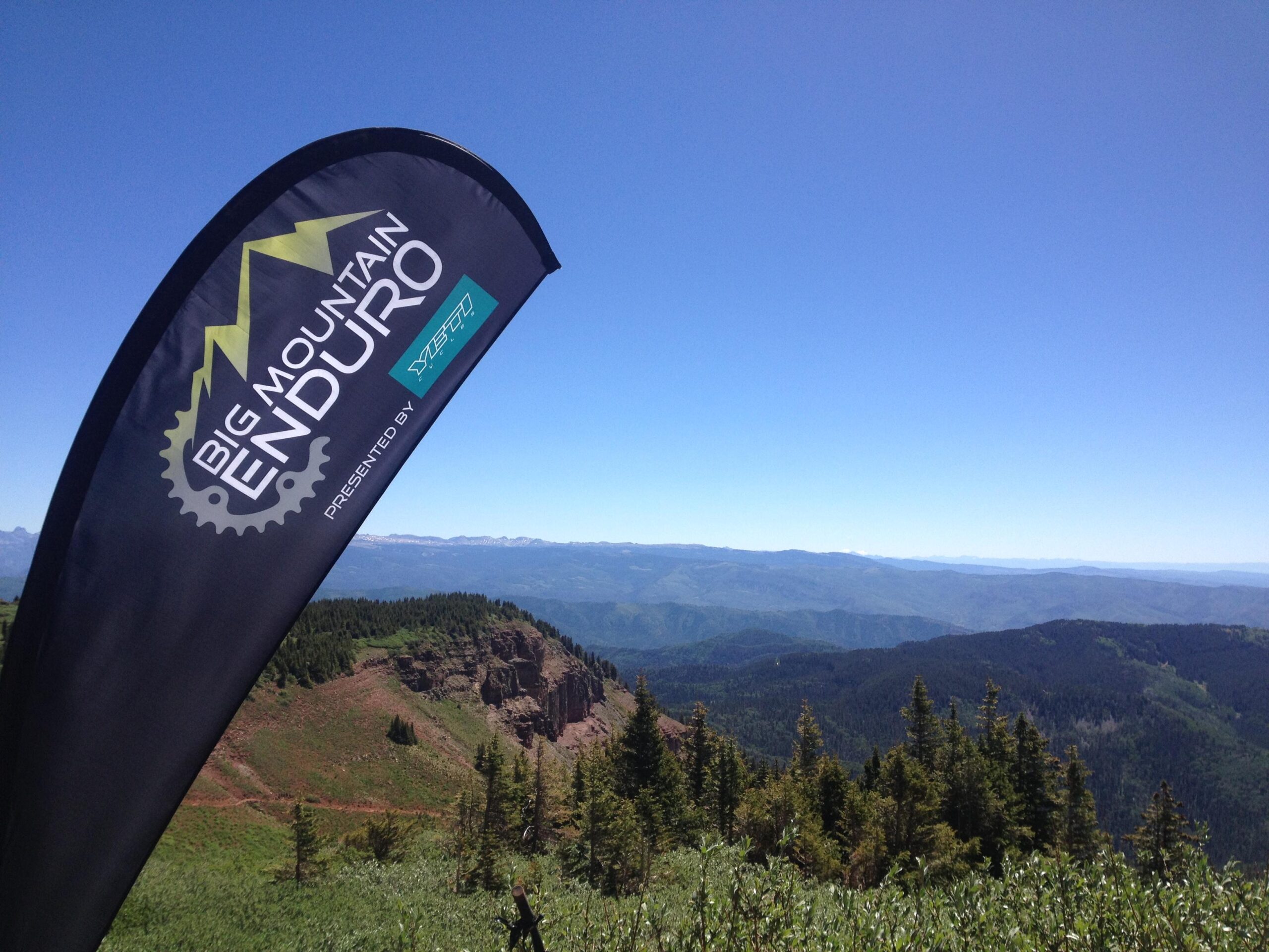 A flag for the Big Mountain Enduro event stands prominently against a clear blue sky, with a picturesque view of mountains and valleys in the background, showcasing lush greenery and distant peaks. Colorado Trail: Kennebec Pass To Junction Creek mountain bike trail.