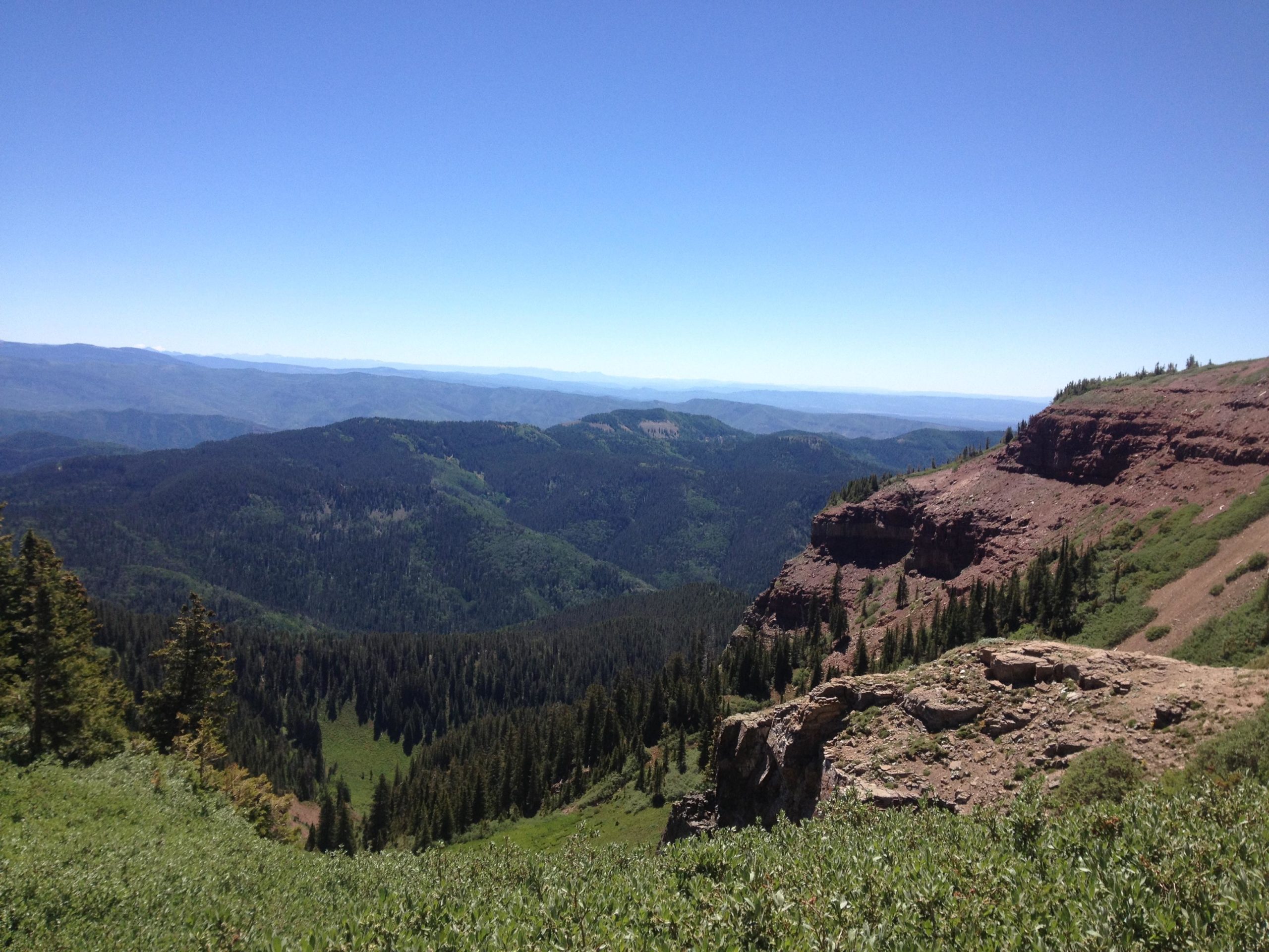 A panoramic view of a mountainous landscape under a clear blue sky. The image features rolling green hills covered in dense forests, rocky outcrops, and distant mountain ranges fading into the horizon. The foreground includes a rugged cliffside with patches of vegetation. Colorado Trail: Kennebec Pass To Junction Creek mountain bike trail.