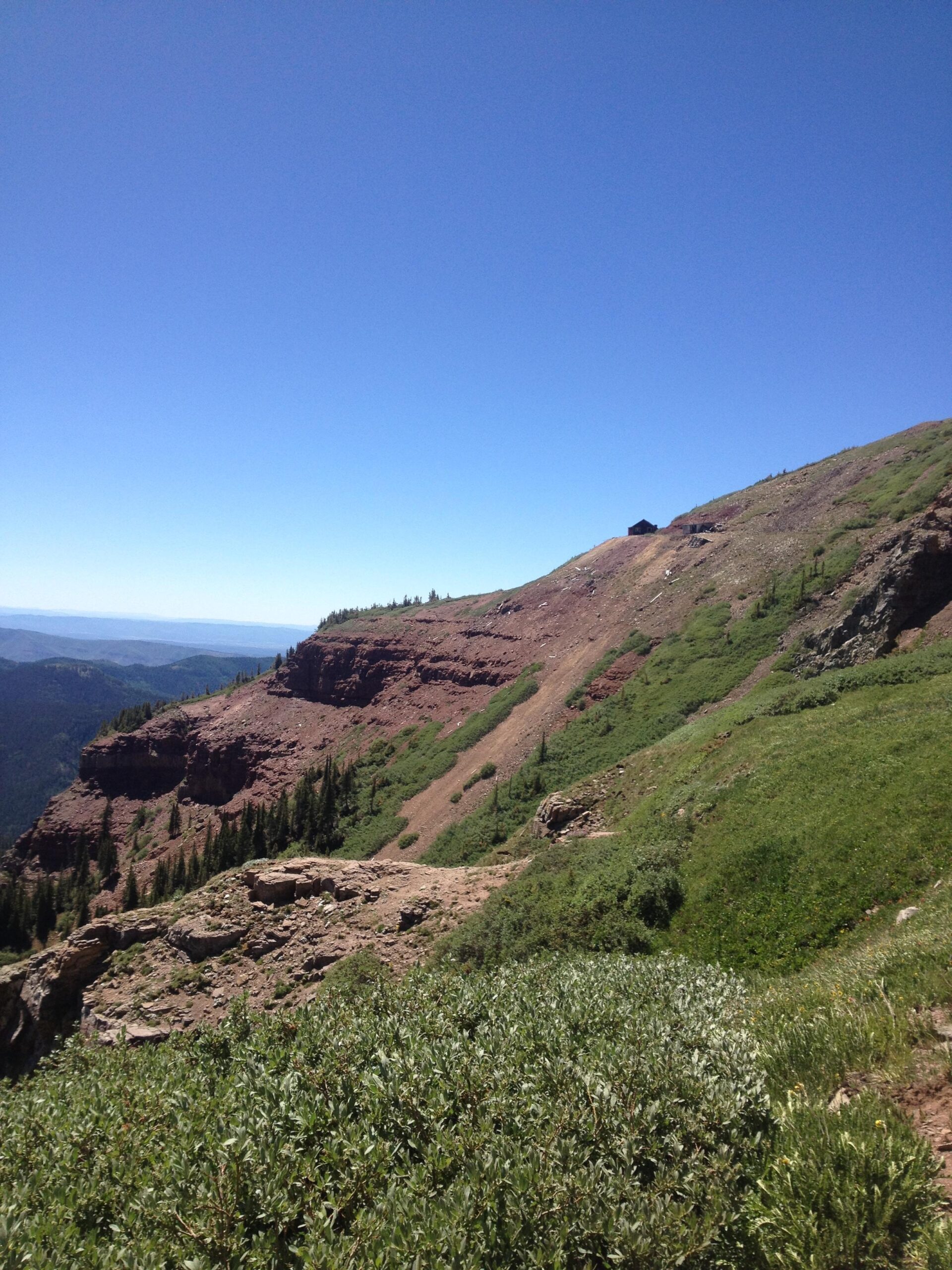 A scenic view of a mountainous landscape under a clear blue sky, featuring a mix of rocky terrain and lush greenery. The slope of the mountain is visible in the foreground, gradually rising towards a rocky outcrop in the distance. Sparse coniferous trees dot the hillside, providing a natural contrast to the reddish-brown earth. Colorado Trail: Kennebec Pass To Junction Creek mountain bike trail.