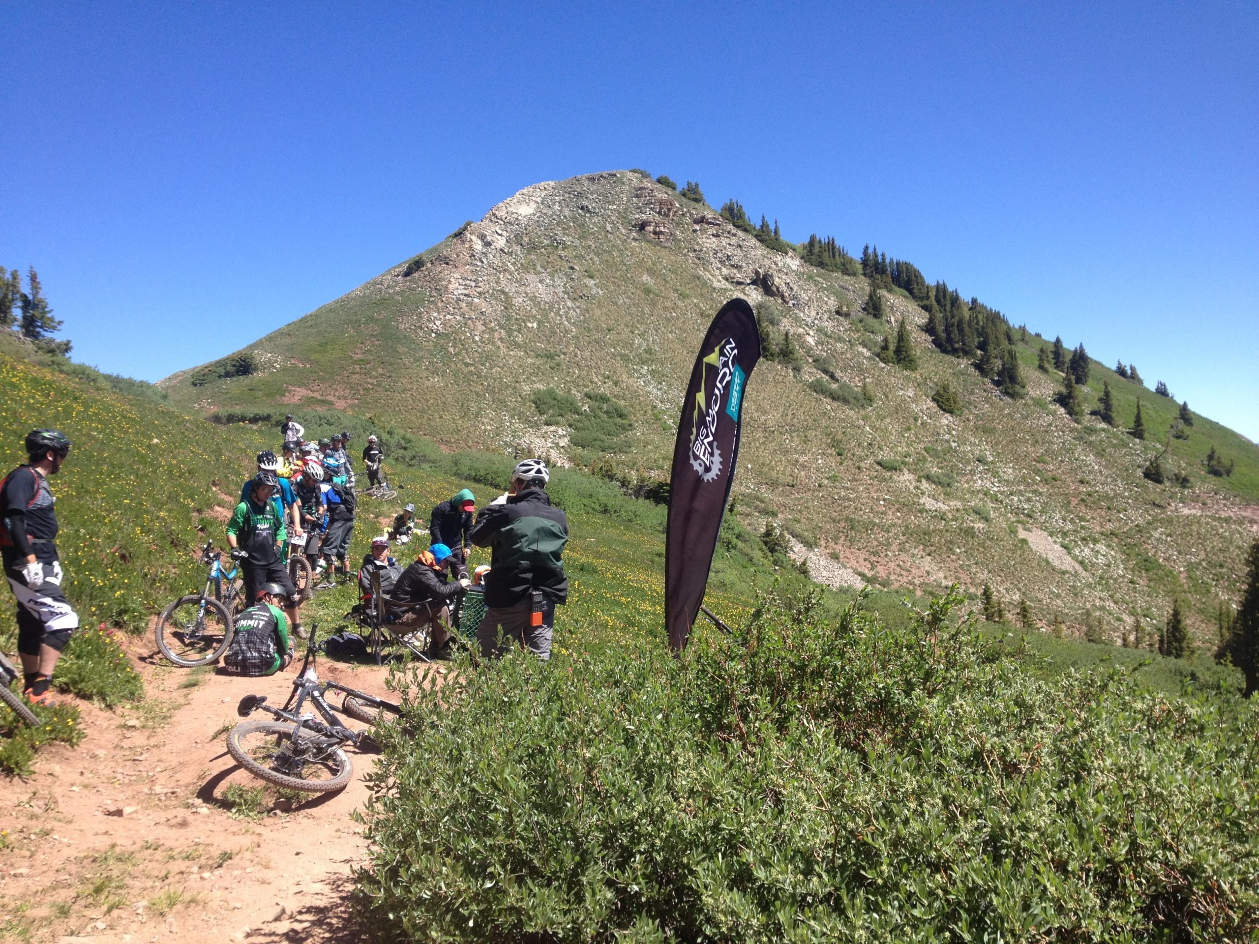 A group of mountain bikers gather on a dirt trail surrounded by lush green hills and a clear blue sky. Some cyclists are resting, while others are engaged in conversation near a flag displaying a mountain biking logo. Bright wildflowers dot the landscape, and a rocky slope rises in the background. Colorado Trail: Kennebec Pass To Junction Creek mountain bike trail.