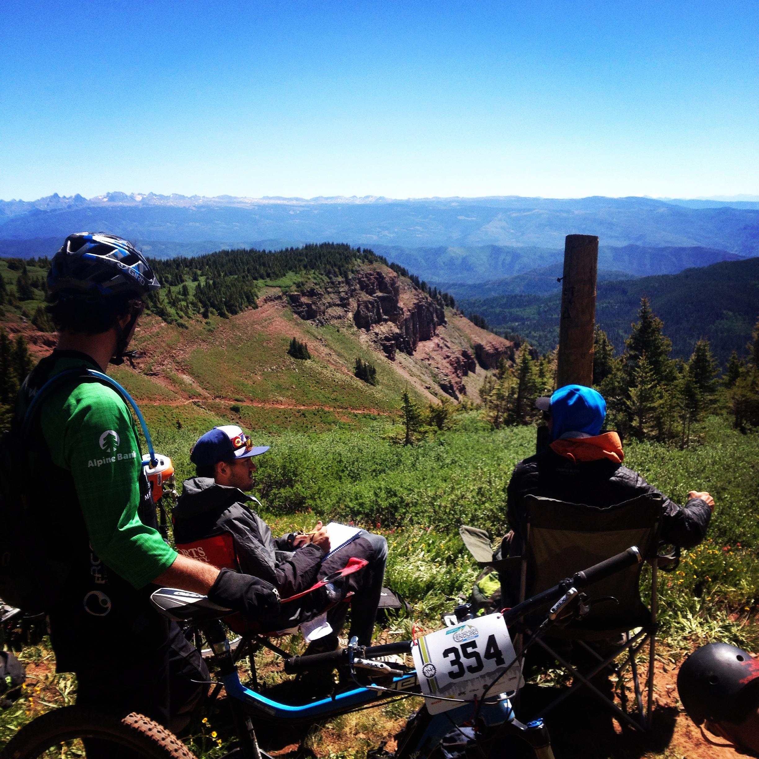 Three mountain bikers enjoy a scenic view from a ridge, surrounded by lush greenery and mountains in the background. One rider stands with their back to the camera, wearing a helmet and a green shirt, while two others sit in folding chairs, one in a blue hoodie and the other dressed more casually. A bike with a race number, 354, is positioned nearby, indicating a mountain biking event or competition. The sky is clear and blue, marking a bright, sunny day. Colorado Trail: Kennebec Pass To Junction Creek mountain bike trail.