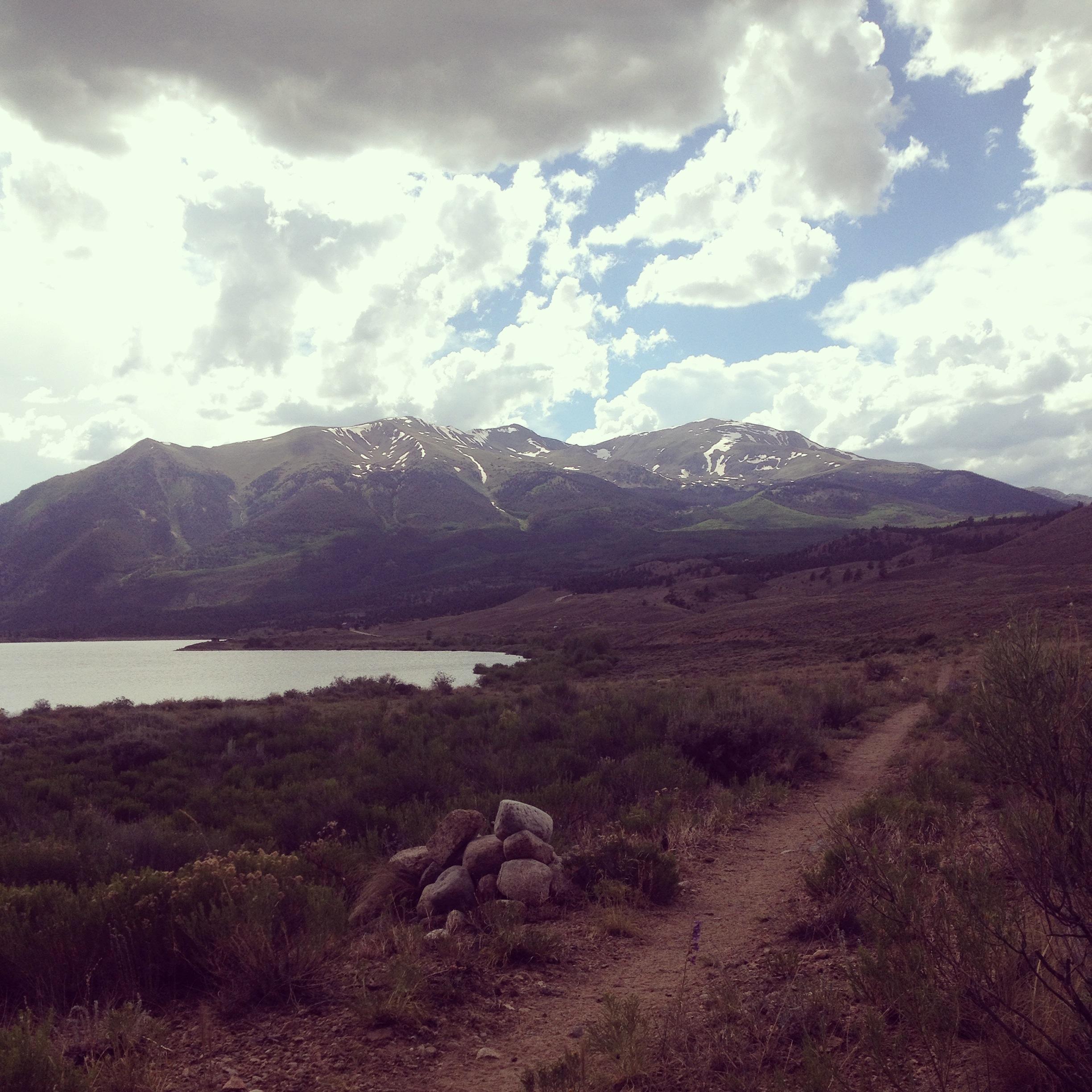A scenic landscape featuring mountains in the background, partially covered in snow, with a calm lake on the left. The foreground includes a rocky outcrop and a dirt path bordered by greenery under a partly cloudy sky. Colorado Trail: Clear Creek Thd to Lake View CG / Hwy 82 mountain bike trail.