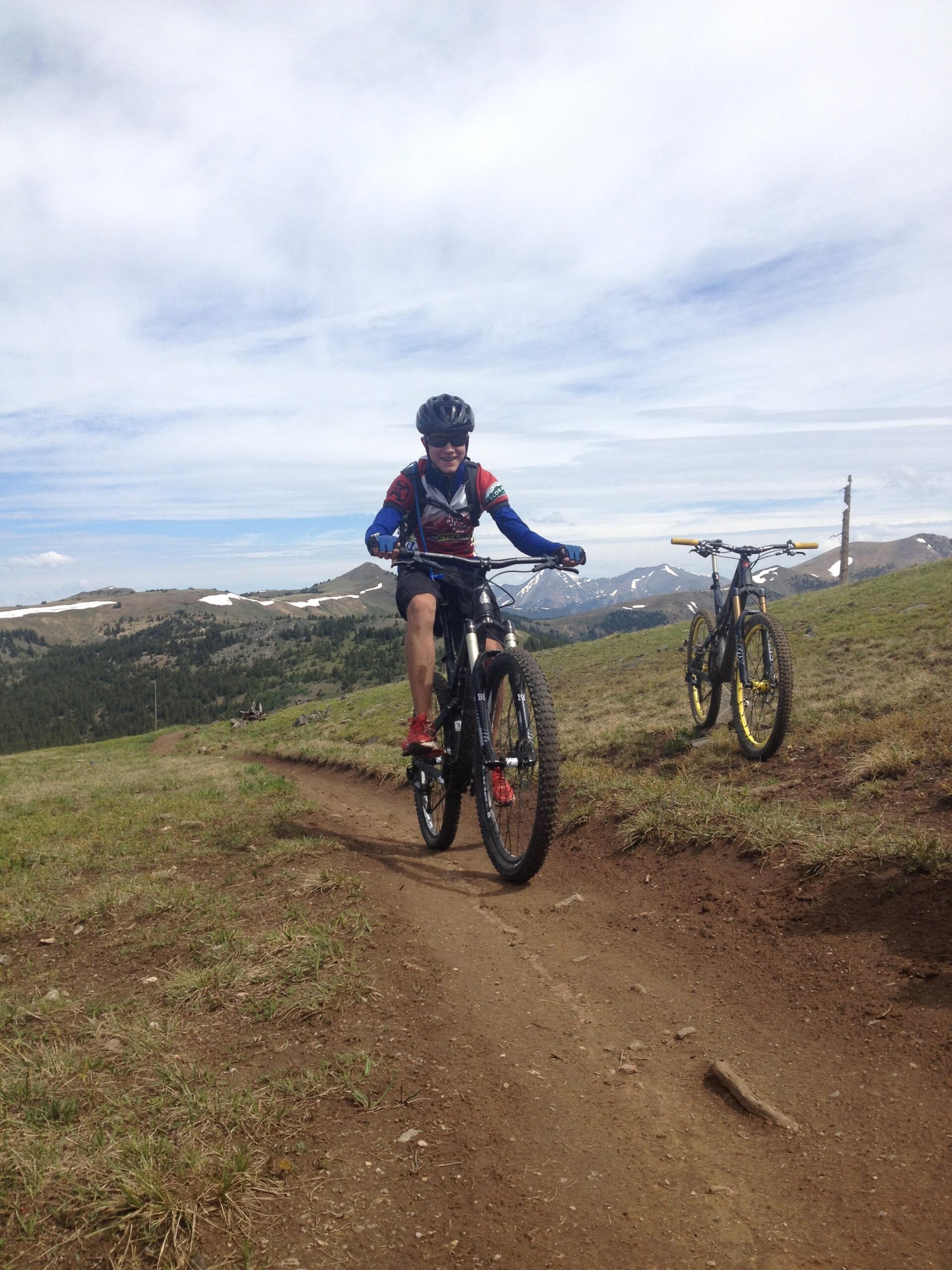 A mountain biker riding along a dirt trail in a grassy landscape, with mountains and clouds in the background. Another bike is parked nearby. The cyclist is wearing a helmet, sunglasses, and a colorful jersey, smiling as they navigate the terrain. Monarch Crest Trail mountain bike trail.
