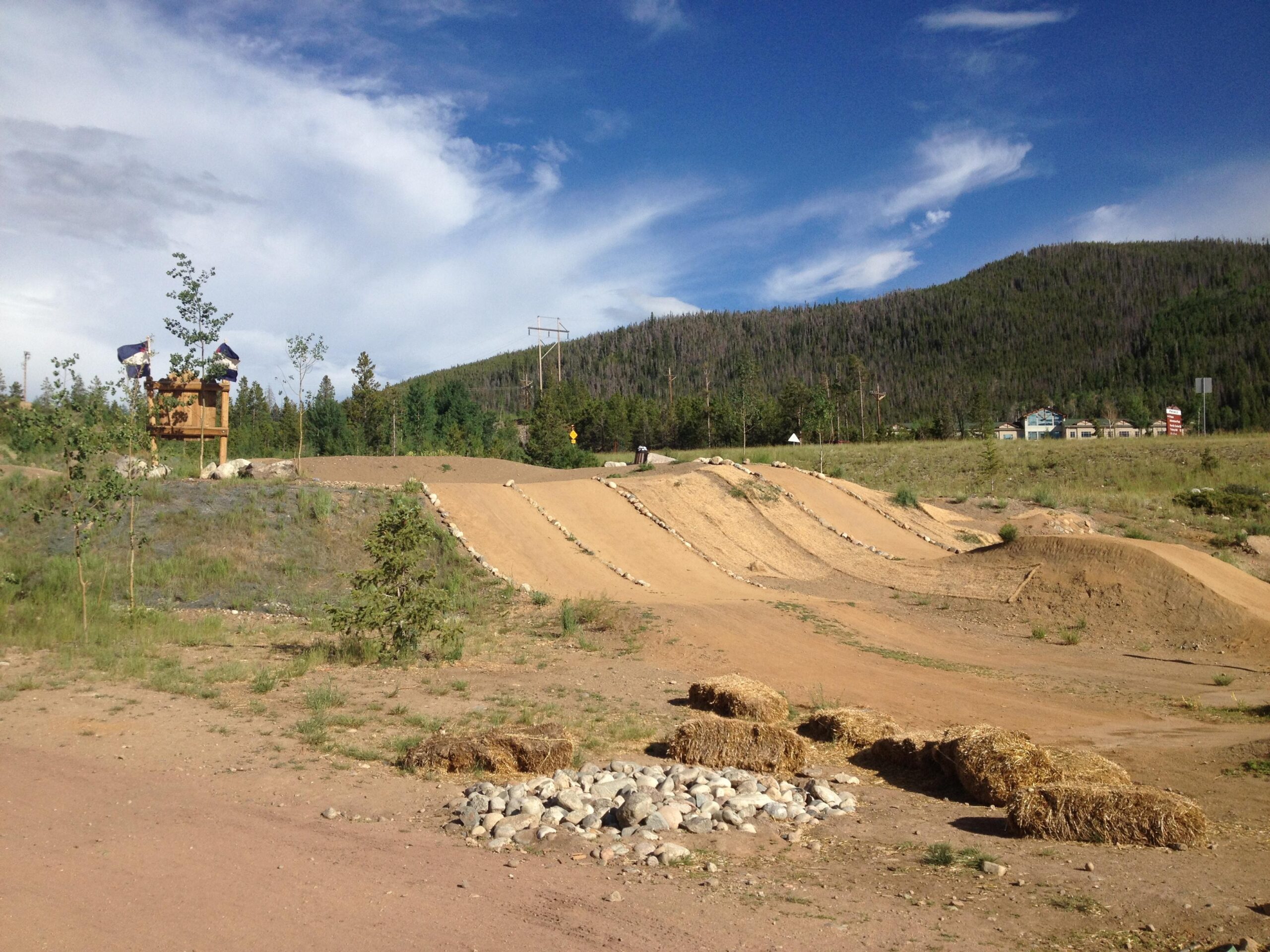 A dirt bike track featuring a series of ramps and jumps, surrounded by green grass and small trees. In the background, a wooden structure with flags is visible, along with a mountain landscape under a partly cloudy sky. Bales of hay and stones are placed along the track. Frisco Adventure Park mountain bike trail.