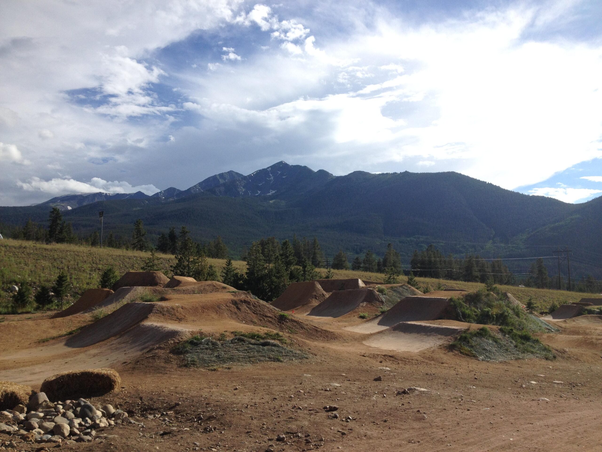 A dirt bike park featuring a series of dirt jumps and ramps, set against a backdrop of lush green mountains and a partly cloudy sky. The foreground shows several jumps, while the mountains rise majestically in the distance under a light blue sky. Frisco Adventure Park mountain bike trail.