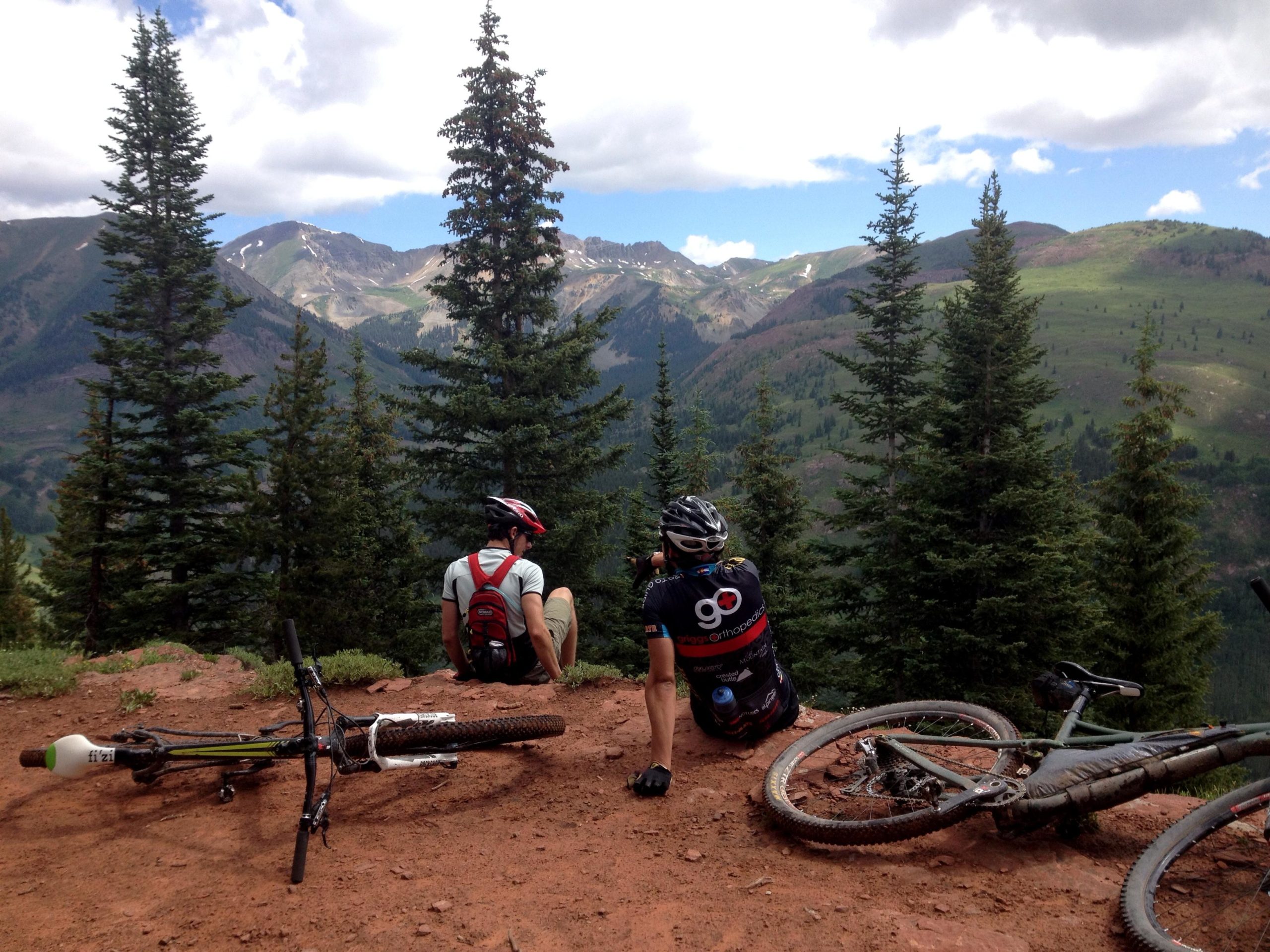 Two mountain bikers resting on a rocky outcrop with a panoramic view of green mountains and trees. One rider sits on the ground, while the other leans back, both bikes nearby. The sky is partly cloudy, creating a serene outdoor atmosphere. Teocalli Ridge mountain bike trail.