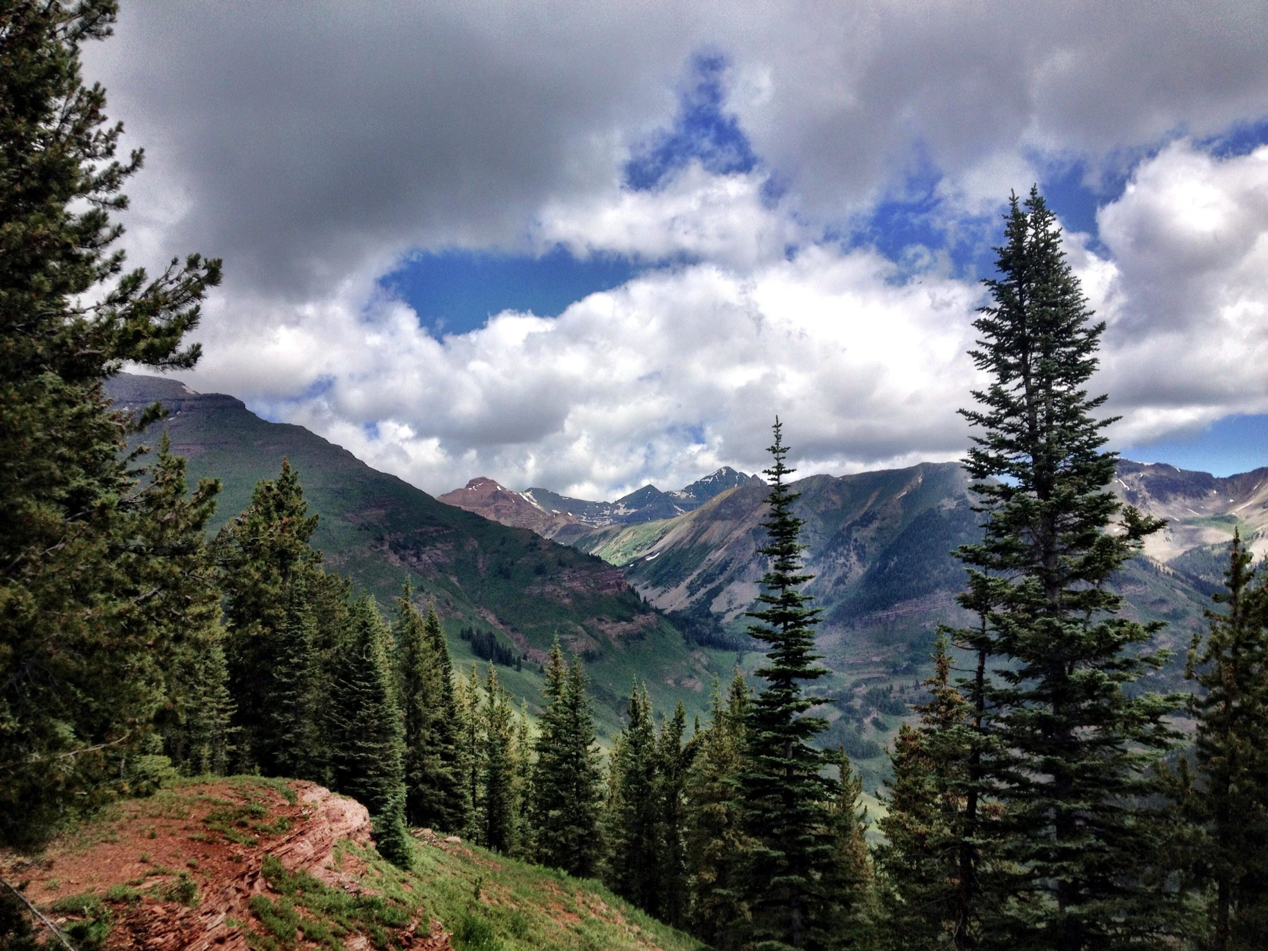 A scenic view of a mountainous landscape featuring lush green hills and tall trees, under a partly cloudy sky. The image captures the natural beauty of the outdoors, showcasing the rolling mountains and a hint of snow-capped peaks in the background. Teocalli Ridge mountain bike trail.