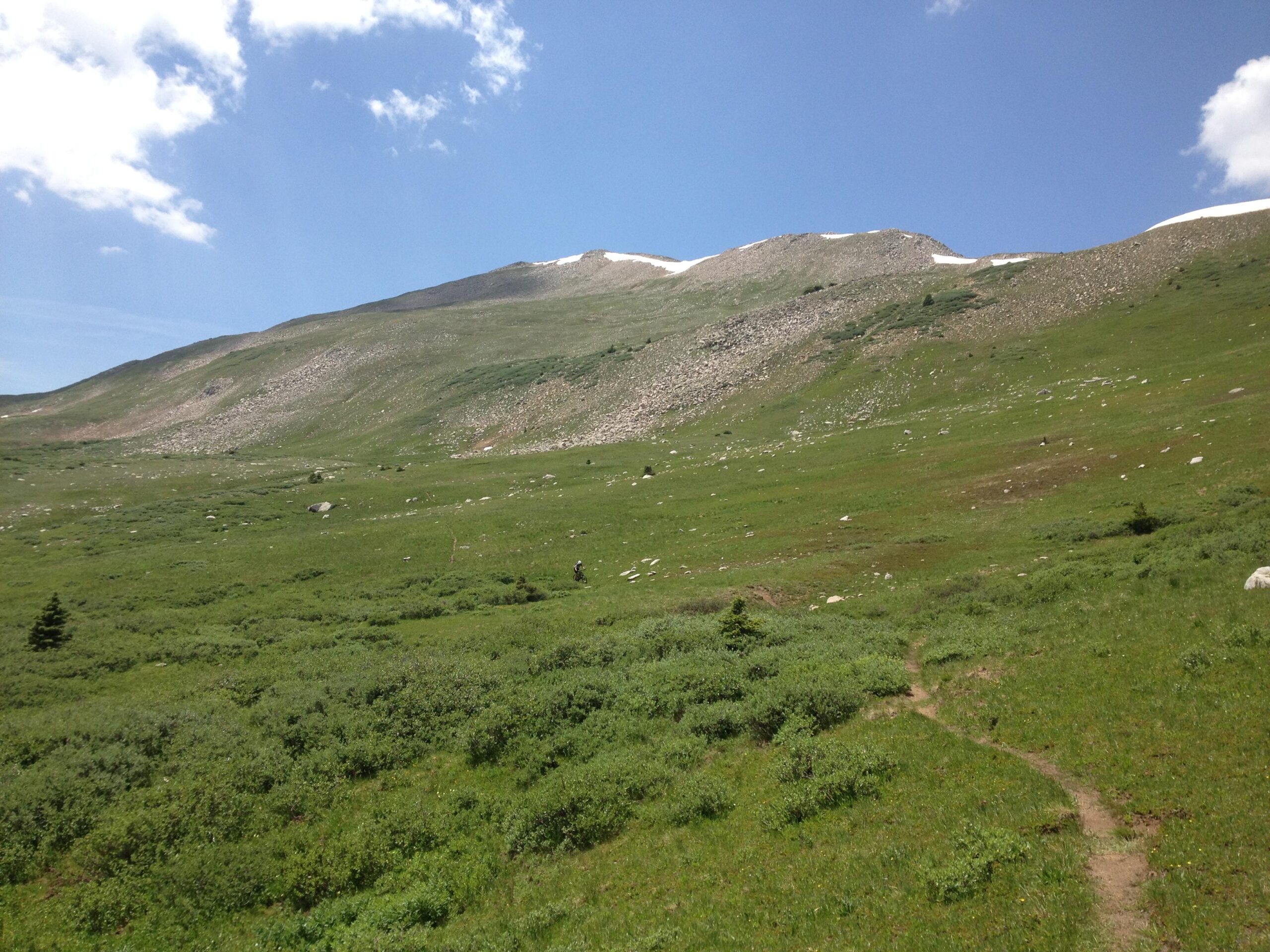 A scenic mountain landscape featuring a gentle sloping hillside covered in lush green grass and scattered boulders. In the distance, a rocky mountain peak is visible beneath a partly cloudy blue sky. A narrow dirt path meanders through the foreground, inviting exploration of the natural terrain. CDT: Alpine Tunnel mountain bike trail.