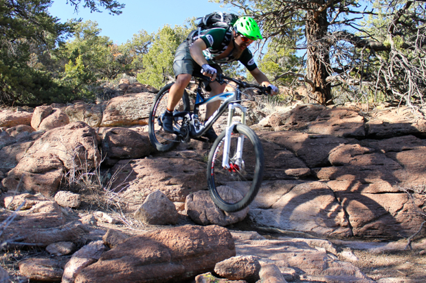 A mountain biker in a green helmet navigates rocky terrain, jumping over boulders in a wooded area. Sunlight filters through the trees, illuminating the path ahead.
