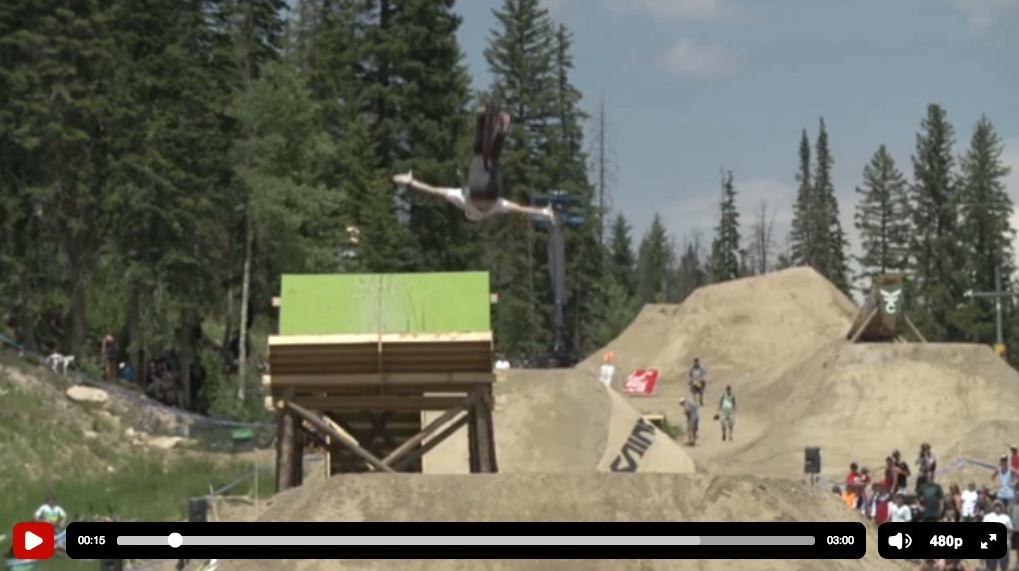 A skier performing a mid-air stunt above a large jump, with a backdrop of trees and spectators in the distance. The scene captures the excitement of an outdoor winter sports event.