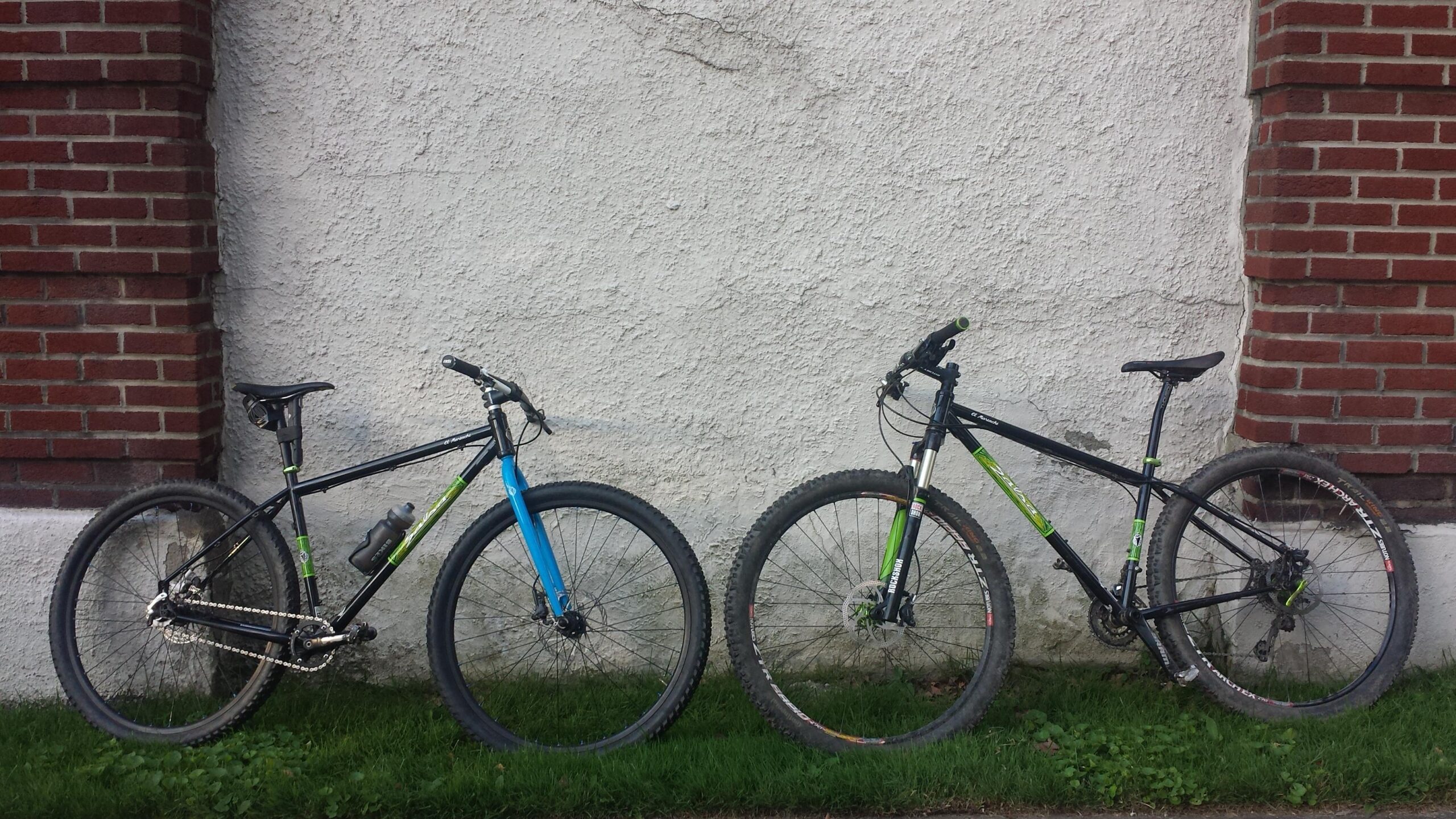 Two mountain bikes are parked side by side against a textured white wall. The bike on the left has a blue front fork and green accents, while the bike on the right features a black frame with green detailing. Both bikes have knobby tires, indicating they are designed for off-road use. The ground is grassy, and there is a red brick column visible on the left side of the image. Frick Park mountain bike trail.