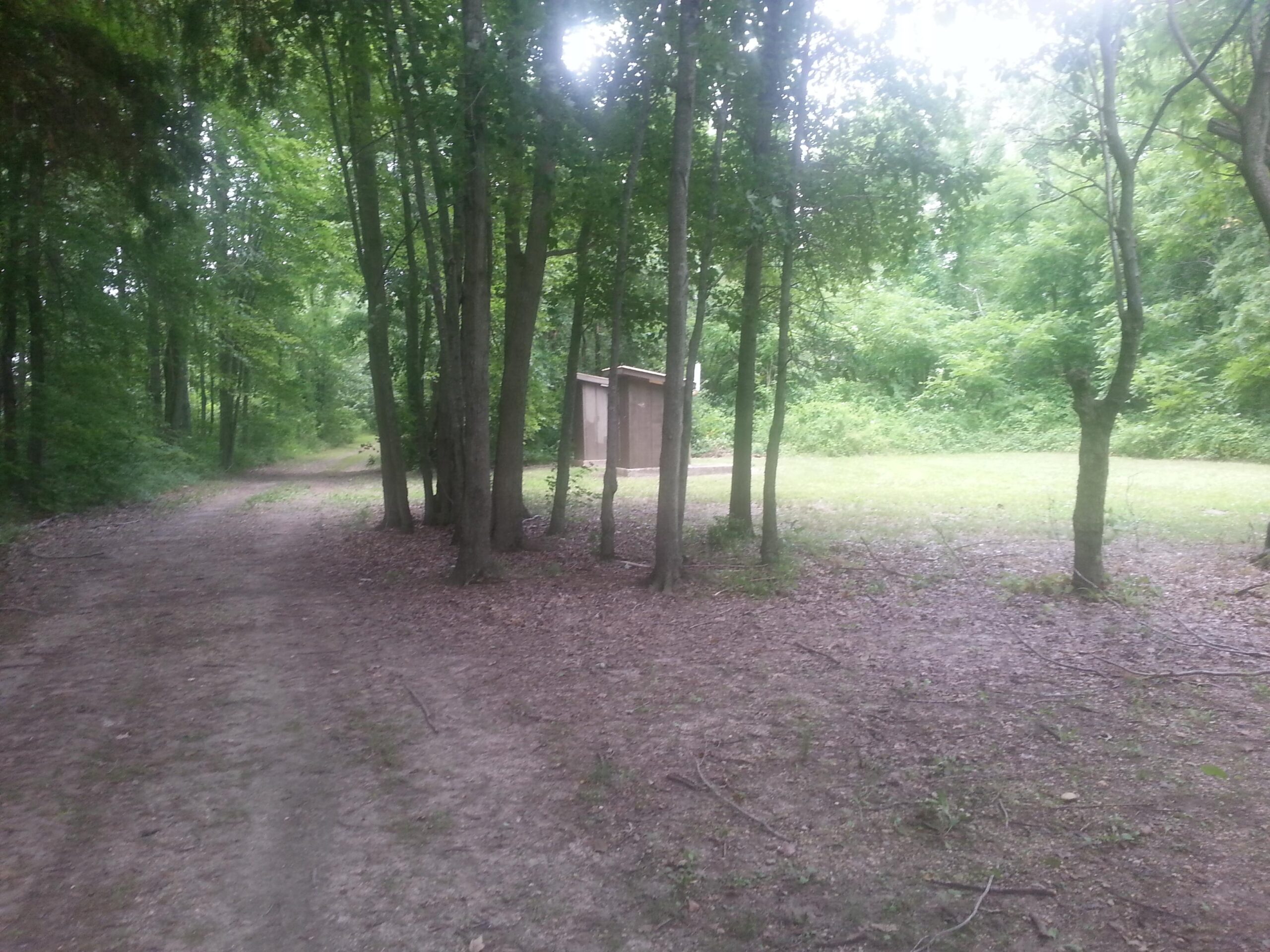 A dirt path winding through a wooded area, flanked by green trees. In the background, a small wooden structure is partially visible amidst the greenery, leading to a patch of open grass. The scene captures a tranquil, natural setting. Newton Lake Equestrian Trail mountain bike trail.