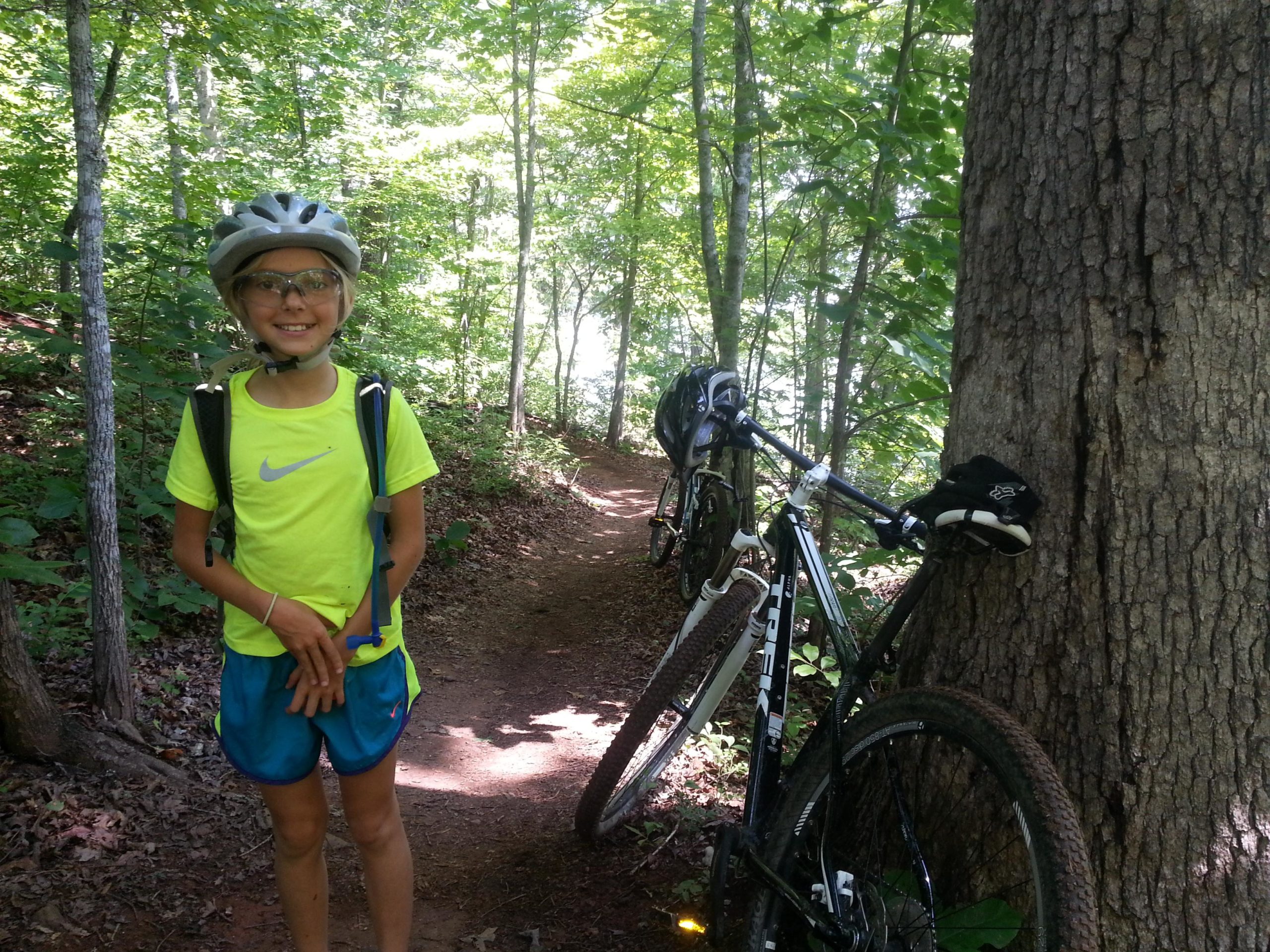 A young child wearing a bright yellow athletic shirt, blue shorts, and protective gear stands on a forest trail, smiling. In the background, a mountain bike leans against a tree, surrounded by lush green foliage. The scene captures a sunny day outdoors, perfect for biking and exploring nature. Jack Rabbit Trails mountain bike trail.