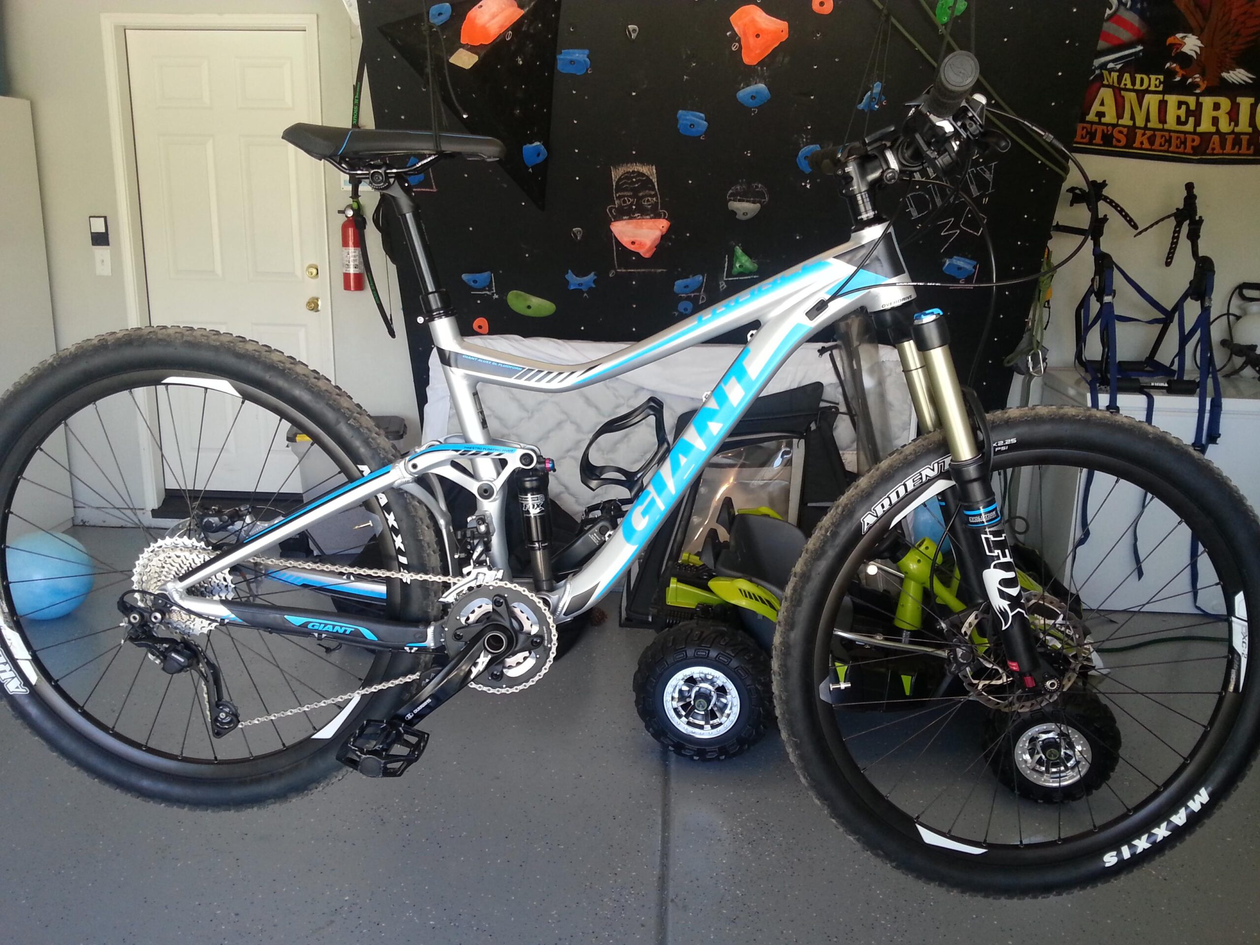 Giant Trance X1: A parked mountain bike with a blue and silver frame, showcasing a suspension system and wide tires, is displayed in a garage setting. The background features a climbing wall and a door, with additional bikes and gear visible.