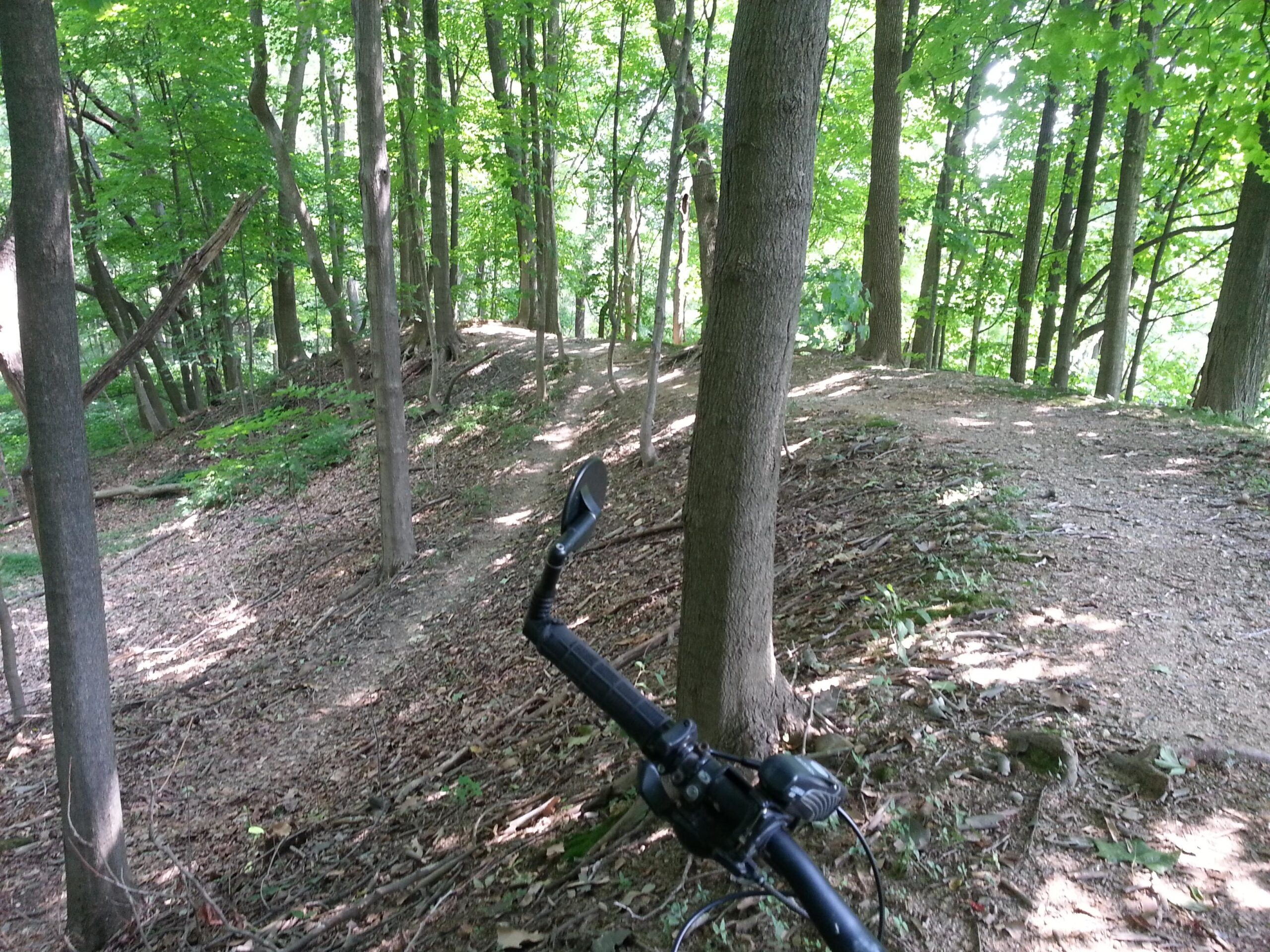 A bike handlebar is visible in the foreground, with a winding dirt trail leading through a lush green forest. Sunlight filters through the trees, illuminating the path that curves around to the right, surrounded by dense foliage and scattered leaves on the ground. Jordan Creek Parkway mountain bike trail.