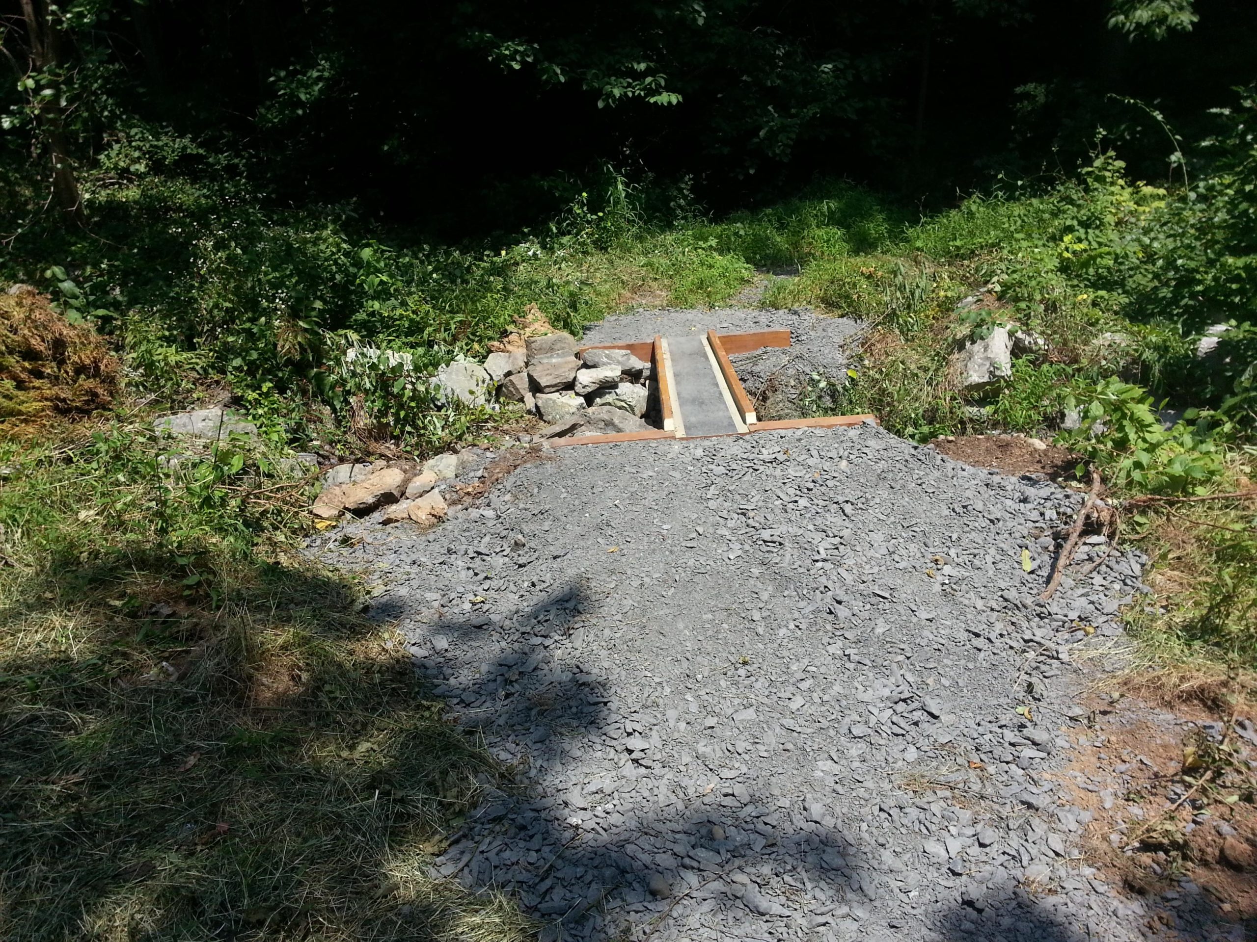 A gravel pathway leading into a wooded area, featuring a wooden bridge with support beams over a small stream. Surrounding the path are patches of grass, rocks, and greenery, with dappled sunlight filtering through the trees. Jordan Creek Parkway mountain bike trail.