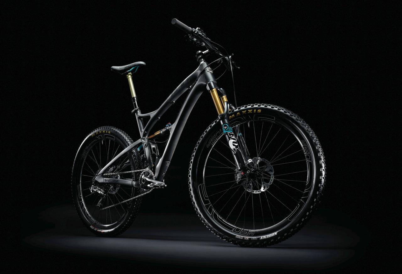 Yeti SB5c: A high-quality mountain bike displayed against a black background, highlighting its sleek design. The bike features a lightweight grey frame with gold suspension forks, black wheels with knobby tires, and detailed components like a derailleur and disc brakes. Soft lighting creates a dramatic effect, emphasizing its performance-oriented design.