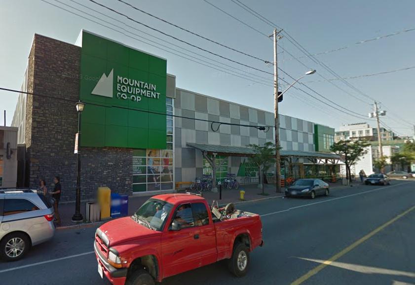 Exterior view of the Mountain Equipment Co-op building, featuring a modern design with a mix of stone and green accents. The entrance is covered by a roof, and there are bicycles parked outside. In the foreground, a red pickup truck and several other vehicles are visible on the street, along with pedestrians and lamp posts.