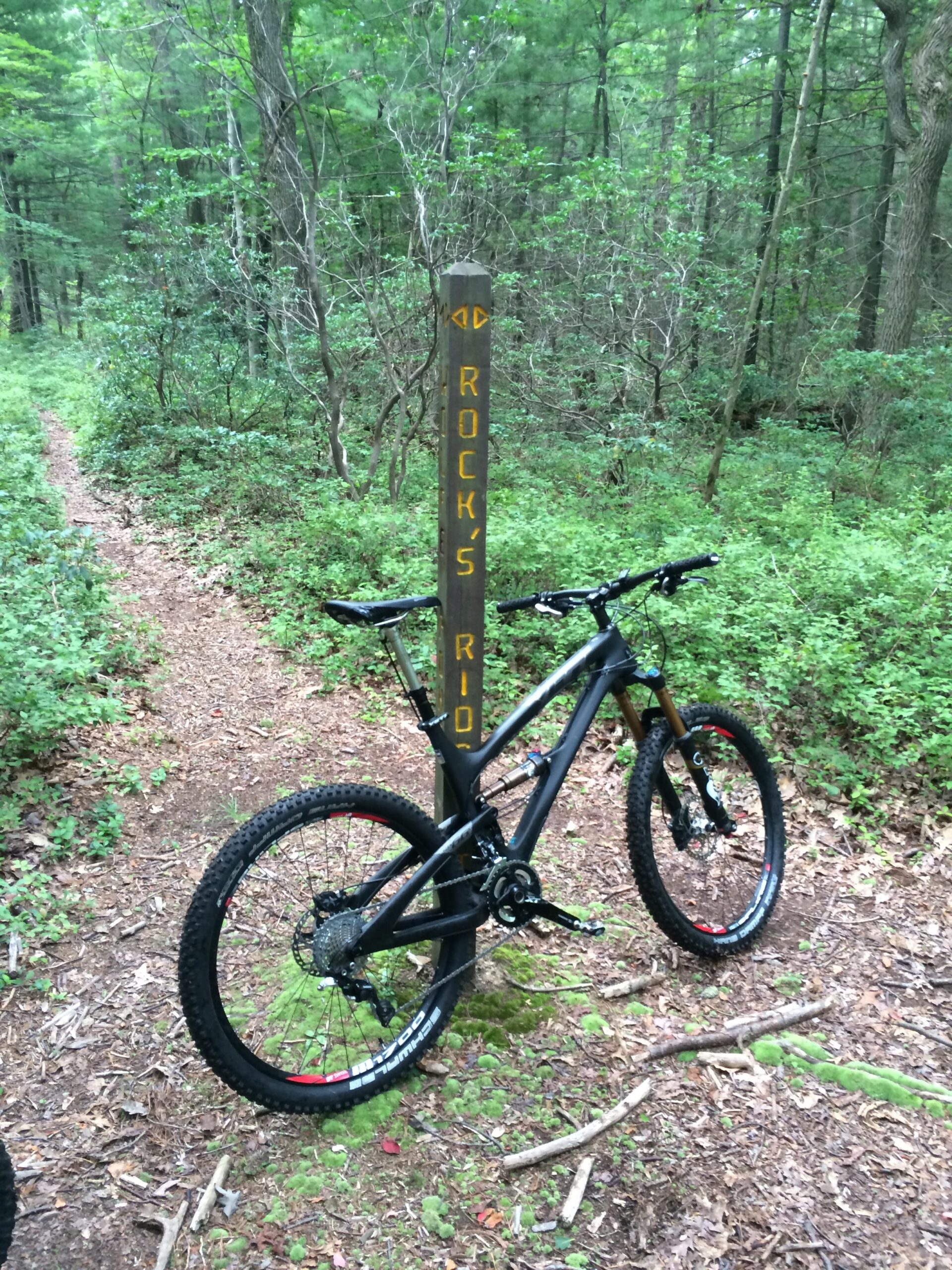 Yeti SB66c: A mountain bike parked beside a wooden trail sign that reads "ROCK'S RD," set in a lush green forest with tall trees and a dirt path visible in the background.