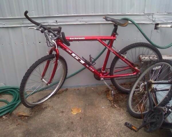 GT Backwoods: A red mountain bike leaning against a wall, positioned next to a partially disassembled bike. The bike features thick tires and a simple handlebar setup, with a garden hose coiled nearby on a concrete surface.