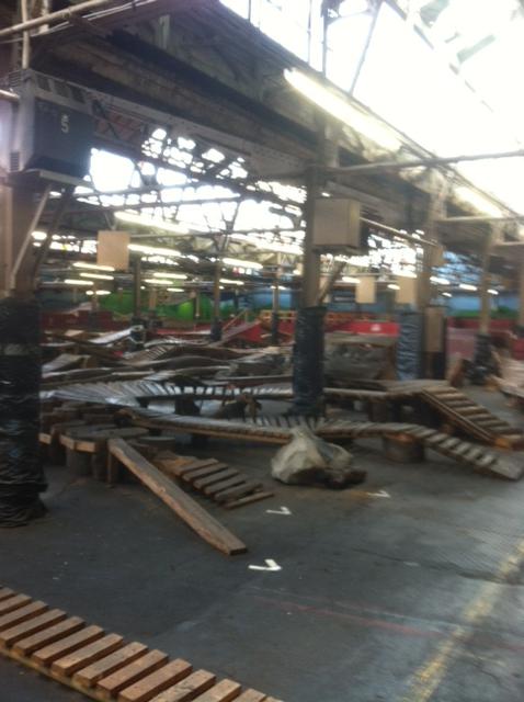 An empty indoor space with a large, open area featuring wooden structures and ramps scattered across the floor. The ceiling shows exposed beams, and sunlight filters through the skylights, illuminating various colored walls in the background. The scene conveys a sense of disarray, with construction materials and debris present. Rays Indoor Mtb Park mountain bike trail.