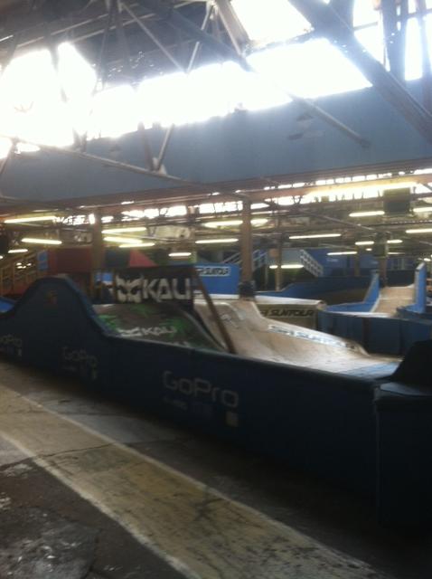 Indoor skatepark featuring ramps and obstacle courses, with various surfaces for skateboarding and biking. The space is illuminated by natural light from skylights and has colorful signage visible on the walls and structures. Rays Indoor Mtb Park mountain bike trail.