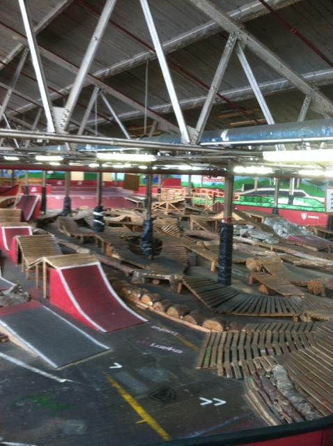 An indoor skate park featuring various ramps, wooden structures, and terrain obstacles for skateboarding and BMX biking, with a high ceiling and vibrant wall art in the background. Rays Indoor Mtb Park mountain bike trail.