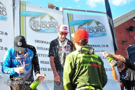 A group of competitors celebrates on the podium at the Big Mountain Enduro event, with champagne being sprayed as they enjoy their victory. The backdrop features a banner with the event name and locations. The scene is lively and filled with excitement, capturing the spirit of competitive mountain biking. Keystone Resort Bike Park mountain bike trail.