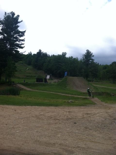 A gravel path leads through a grassy area with trees in the background. In the distance, a dirt ramp is visible, likely used for biking or jumping. A cyclist in a green outfit is riding along the path near the foreground. The sky is overcast, hinting at a cloudy day. Highland Mountain Bike Park mountain bike trail.