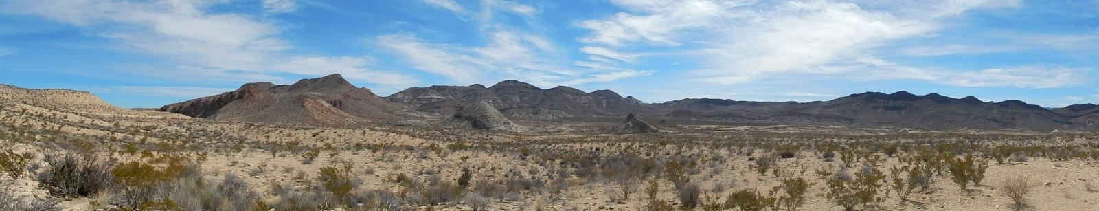 A panoramic view of a desert landscape showcasing rolling hills and mountains under a blue sky with wispy clouds. The foreground features sparse vegetation, including low shrubs and grasses, highlighting the arid environment. Big Bend State Park mountain bike trail.
