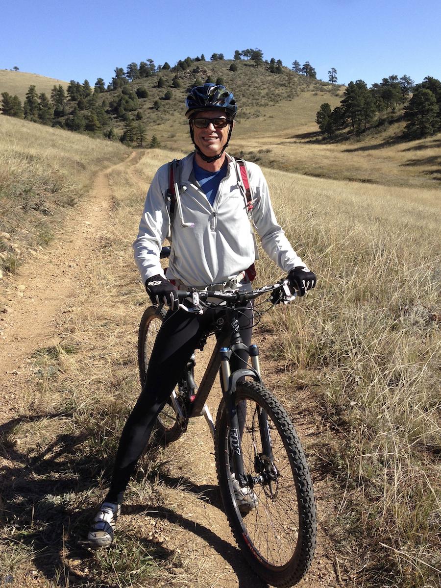 Pivot Mach 429: A smiling cyclist wearing a helmet and sunglasses stands beside a mountain bike on a dirt trail. The background features rolling hills and scattered trees under a clear blue sky. The cyclist is dressed in a light jacket and tight pants, ready for an outdoor adventure.